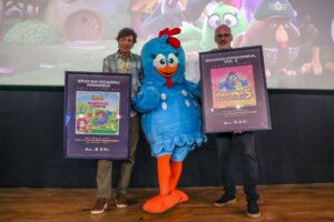 Personagem Galinha Pintadinha ao lado de participantes com materiais do evento